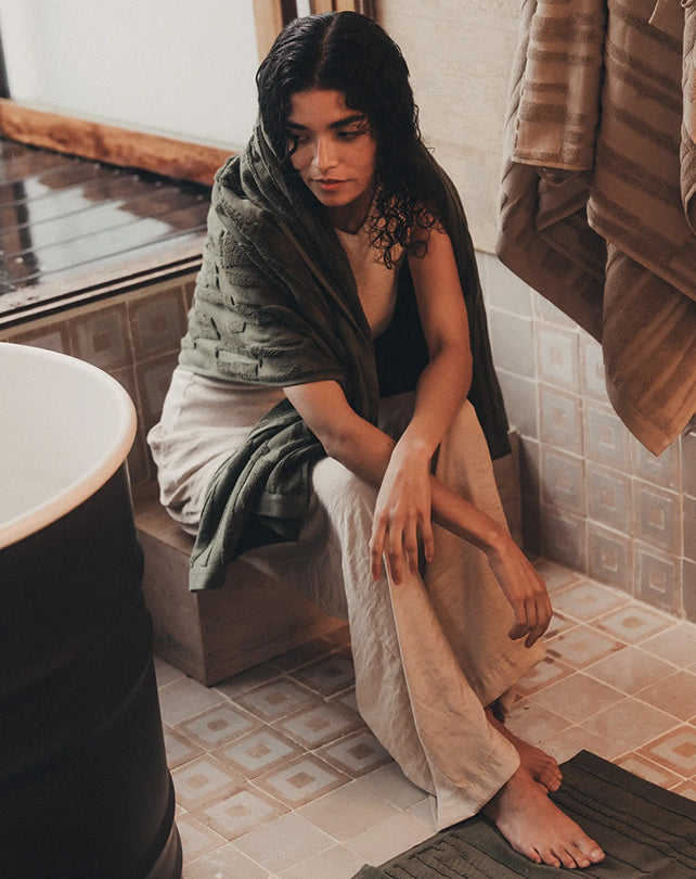 Woman wrapped in a towel sitting on a bath mat in a bathroom.