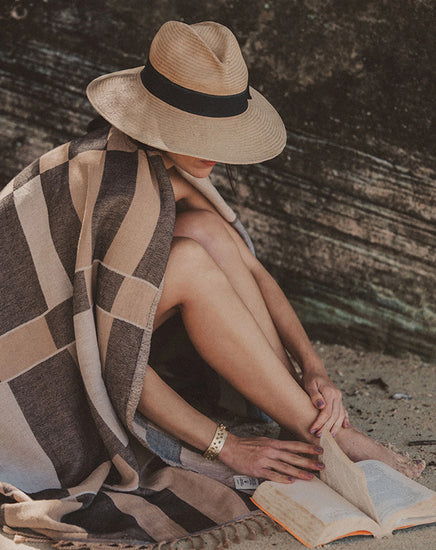 Person sitting on a beach with a hat, towel, and book