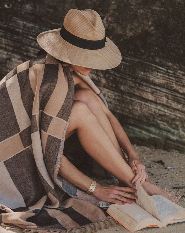 Person sitting on a beach with a hat, towel, and book