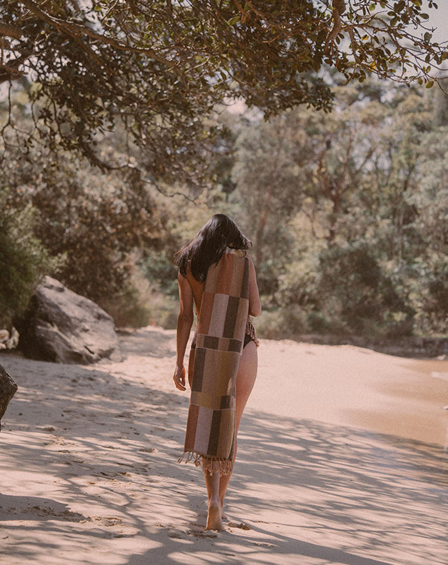 Woman walking on a sandy path with a patterned towel draped over her shoulder, surrounded by trees and nature.