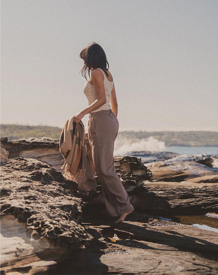 Person standing on a rocky beach holding a bag, with a scenic view of water and sky.