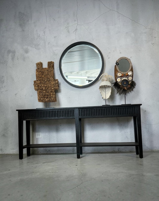 A black wooden console table against a textured gray concrete wall, styled with a round black-framed mirror, a carved wooden sculpture, a white hat on a stand, and a decorative mask.