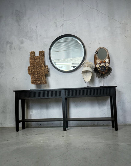 A black wooden console table against a textured gray concrete wall, styled with a round black-framed mirror, a carved wooden sculpture, a white hat on a stand, and a decorative mask.