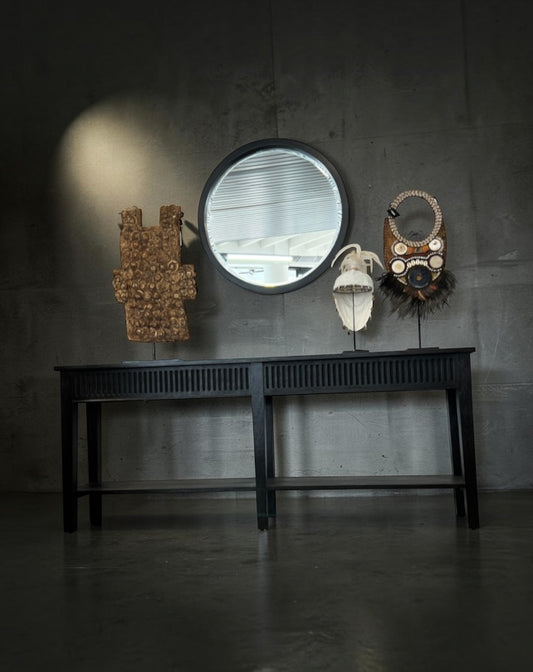 A black console table against a dark concrete wall, styled with a round mirror, a carved wooden sculpture, a white skull, and a decorative mask.