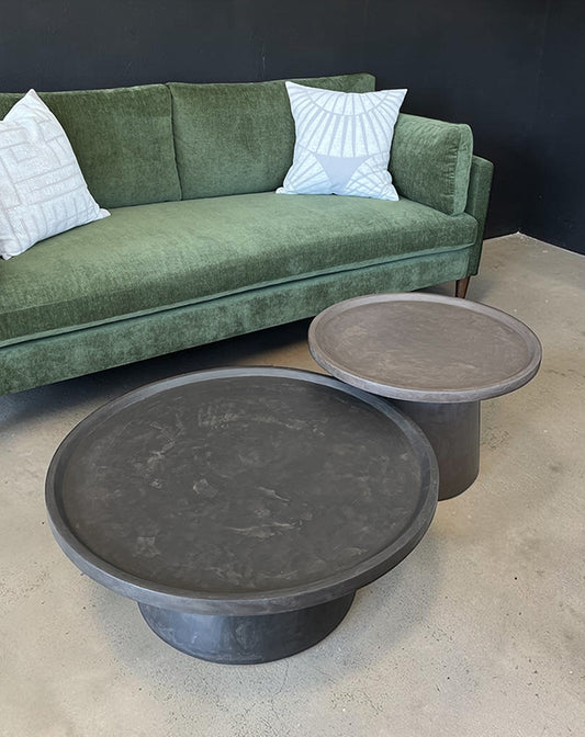 Two dark gray coffee tables in front of a green sofa with white pillows.