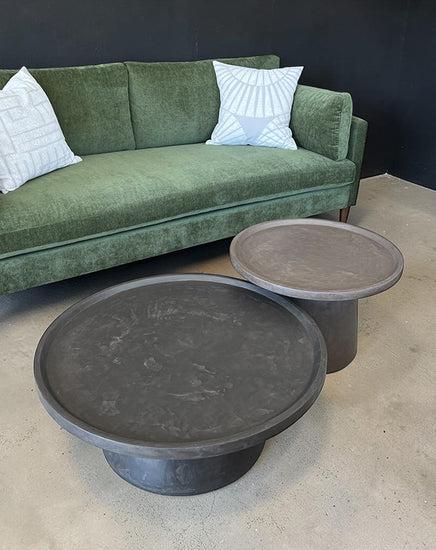 Two dark gray coffee tables in front of a green sofa with white pillows.